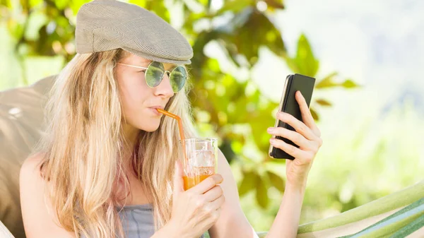 用智能手机对着金发碧眼的女人微笑 在花园里的吊床上放松一下 喝点果汁 利用空闲时间和暑假的概念上网或通过社交媒体与朋友聊天 — 图库照片