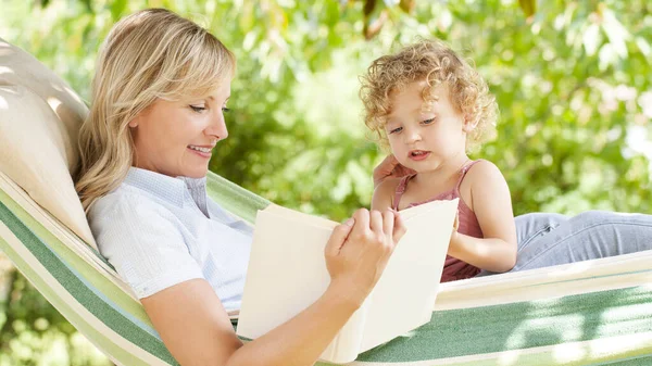 smiling mom reading a story from the book to the little girl daughter child blue eyes with blond curly hair, together lying on the hammock in the green home garden, lovely family concept