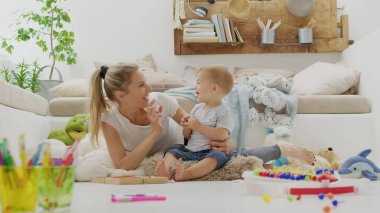 Mutlu gülümseyen anne bebeğiyle oynuyor, renkli kalemler ve tebeşirlerle, eğitici öğrenme oyunlarıyla, yastıklarla hayvanlar arasında oturarak, pelüş, sağlıklı ve büyüme konseptiyle ilgilenerek. 