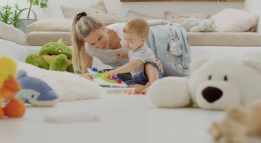 Mutlu gülümseyen anne, renkli eğitim oyuncağıyla güzel bebeğiyle oynuyor. Yastıklar ve hayvanların arasında oturuyor. Rahat, sağlıklı ve büyüme konseptiyle ilgileniyor.