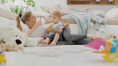 Mutlu gülümseyen anne bebeğiyle oynuyor, renkli kalemler, keçeli kalemler ve beyaz tahta ile, eğitici öğrenme oyunu, yastıklar ve hayvanlar arasında evde oturmak, pelüş, sağlıklı ve büyüme konseptiyle ilgilenmek. 