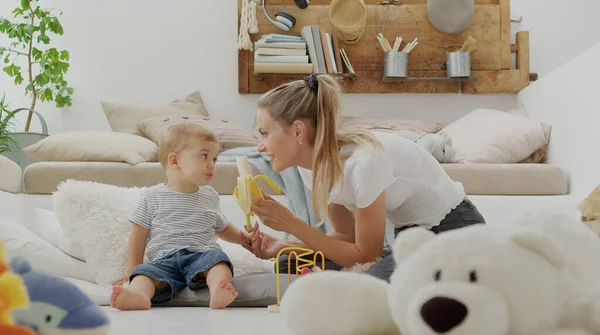 Mutlu ve gülümseyen bir anne bebeğinize vitamin ve mineral bakımından zengin, yastıklar ve pelüş oyuncaklar arasında evde oturan sağlıklı ve büyüme konseptiyle ilgilenen dengeli bir diyet için muz yediriyor.