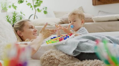 Mutlu gülümseyen anne güzel bebeğiyle oynuyor, renkli eğitim oyuncağıyla, evde yastıkların arasında yatıyor, sağlıklı ve büyüme konseptiyle ilgileniyor.