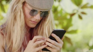 Güneş gözlüklü gülümseyen sarışın kadın akıllı telefon kullanıyor, bahçedeki hamakta uzanıyor, internette sörf yapmak veya sosyal medyayı kullanarak arkadaşlarıyla sohbet etmek için boş zaman ve yaz tatili konsepti.