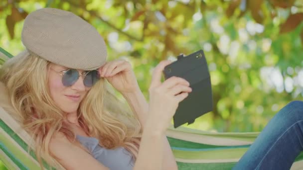 スマートフォンを使用してサングラスをかけブロンドの女性を笑顔 庭のハンモックでリラックスして横になり 無料の時間と夏休みのコンセプトは インターネットをサーフィンしたり ソーシャルメディアを使用して友人とチャットする — ストック動画