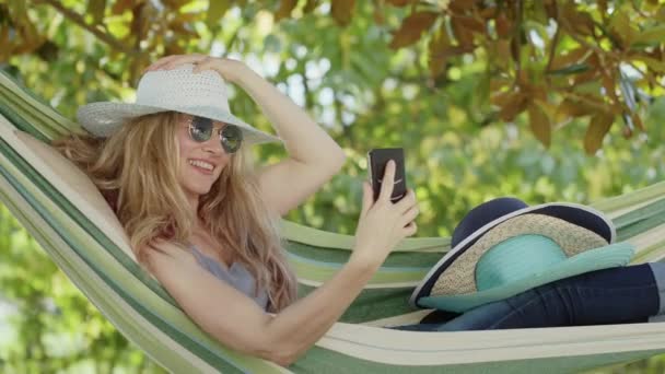 在花园 休闲和夏季的吊床上 戴着太阳镜的金发女人用智能手机微笑着 换了顶帽子 制作了一部具有挑战性的视频 并在网上或社交媒体上发表了自己的观点 — 图库视频影像