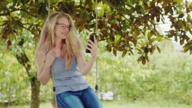 Gözlüklü gülümseyen, akıllı telefon kullanan, salıncakta sallanan, internette sörf yapan ya da sosyal medyayı kullanan arkadaşlarıyla sohbet eden sarışın bir kadın.