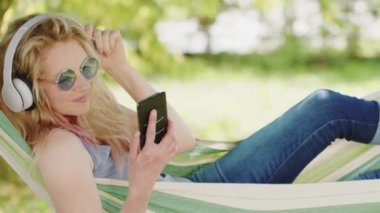 Gülümseyen sarışın kadın akıllı telefon kullanıyor, kulaklıkla müzik dinliyor, bahçedeki hamakta, boş zamanlarında ve yaz konseptinde internet ya da sosyal medya kullanıyor.
