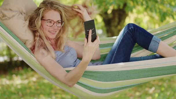 Mujer Rubia Sonriente Con Anteojos Usando Teléfono Inteligente Tumbado Relajándose — Vídeos de Stock
