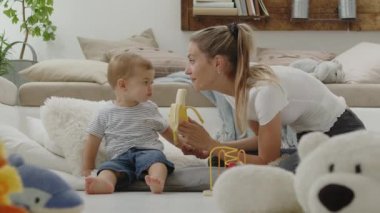 Mutlu ve gülümseyen bir anne bebeğinize vitamin ve mineral bakımından zengin, yastıklar ve pelüş oyuncaklar arasında evde oturan sağlıklı ve büyüme konseptiyle ilgilenen dengeli bir diyet için muz yediriyor.