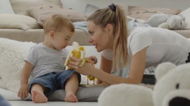 Mutlu ve gülümseyen bir anne bebeğinize vitamin ve mineral bakımından zengin, yastıklar ve pelüş oyuncaklar arasında evde oturan sağlıklı ve büyüme konseptiyle ilgilenen dengeli bir diyet için muz yediriyor.