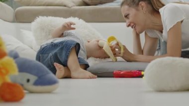 Mutlu ve gülümseyen bir anne bebeğinize vitamin ve mineral bakımından zengin, yastıklar ve pelüş oyuncaklar arasında evde oturan sağlıklı ve büyüme konseptiyle ilgilenen dengeli bir diyet için muz yediriyor.