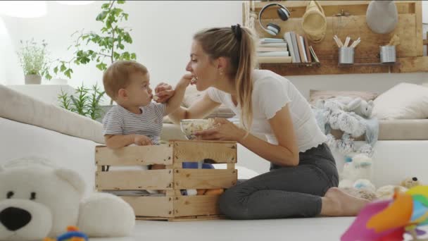 快乐而微笑的妈妈 带着漂亮的孩子坐在木箱里 坐在枕头和毛绒绒的动物中间吃爆米花 健康而又体贴 充满爱心 — 图库视频影像
