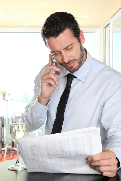 Empresário com celular lendo o jornal, com um copo de vinho — Fotografia de Stock