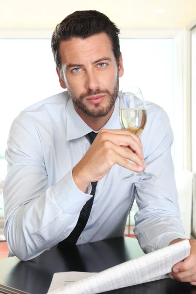 Handsome man drinking a glass of sparkling wine white, sitting at the bar with newspaper — Stock Photo, Image