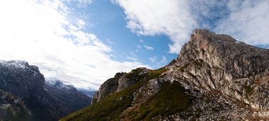 Yüksek dağlık bir manzaranın panoramik manzarası engebeli kayalık tepeler, dağınık bulutlu parlak mavi bir gökyüzü ve sonsuz yeşil ağaçlar, çimenler ve uzak karla kaplı zirveler..