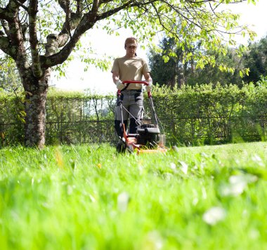 Genç bir adam, çim biçme makinesiyle parlak, yemyeşil bir bahar bahçesi biçiyor. Mevsimlik bahçe bakımı çimenleri sağlıklı ve düzenli tutmaya odaklandı.