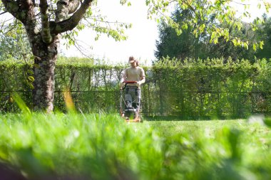 Genç bir adam, çim biçme makinesiyle parlak, yemyeşil bir bahar bahçesi biçiyor. Mevsimlik bahçe bakımı çimenleri sağlıklı ve düzenli tutmaya odaklandı.