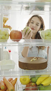 Kadın buzdolabının kapısını açar, mutlu bir şekilde pasta ve kremanın tadına bakar, ama istemeyerek de olsa tatlı ve elma arasında karar verir. Sağlıklı bir diyet seçimi yapıyor. Mutfak dolabının içinden pov çekimi.