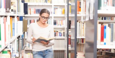 Modern bir kütüphanede kitap okuyan gözlüklü genç bir kadın. Arkaplanda renkli kitaplarla dolu kitaplıklar. Öğrenim, eğitim, öğrenim, bilgi ve akademik yaşam tarzı kavramı
