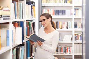 Modern bir kütüphanede kitap okuyan gözlüklü genç bir kadın. Arkaplanda renkli kitaplarla dolu kitaplıklar. Öğrenim, eğitim, öğrenim, bilgi ve akademik yaşam tarzı kavramı