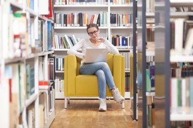 Genç bir üniversite öğrencisi kütüphanede okuyor. O bir koltukta oturuyor, gözlük takıyor ve bir dizüstü bilgisayar kullanıyor. Eğitim, öğrenim, akademik başarı ve sınav hazırlığı kavramı.