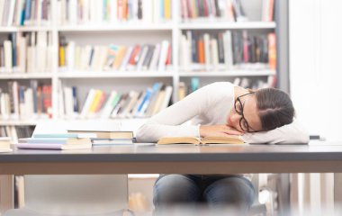 Genç bir üniversite öğrencisi kütüphane masasında uyuyor, ders kitabının üzerinde dinleniyor. Yüksek öğrenimde akademik stres, yorgunluk, aşırı çalışma ve öğrenim hayatı kavramı.