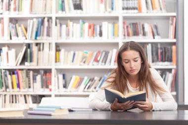 Genç bir üniversite öğrencisi kütüphanedeki masada açık bir kitap okumaya odaklanmış. Öğrenim, öğrenim, konsantrasyon, akademik yaşam ve yüksek öğrenim kavramı.