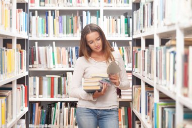 Sırıtkan kız öğrenci kütüphane koridorunda raflarla dolu bir yığın kitap tutuyor. Öğrenim, öğrenim, akademik başarı, konsantrasyon ve eğitimle tatmin kavramı