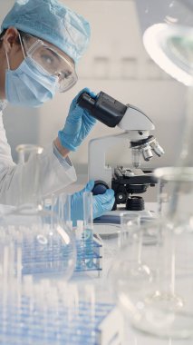 Maske, güvenlik gözlüğü, eldiven ve saç şapkası takan kadın bilim adamı bir laboratuarda mikroskop kullanarak çalışıyor. Mikrobiyoloji, biyoteknoloji, tıbbi ve ilaç araştırmaları.