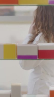 Kadın eczacı, akıllı telefonla konuşurken eczane raflarından doğru ilacı seçiyor. Profesyonel, dikkatli ve güvenilir müşteri hizmetleri sunuyor..