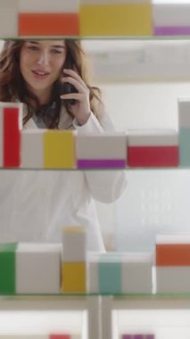 Kadın eczacı, akıllı telefonla konuşurken eczane raflarından doğru ilacı seçiyor. Profesyonel, dikkatli ve güvenilir müşteri hizmetleri sunuyor..