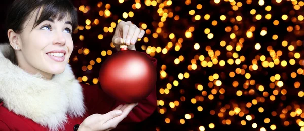 Mujer de Navidad con bola roja, sonriendo y mira hacia arriba en las luces ba — Foto de Stock