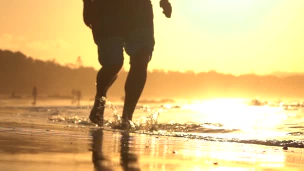 Κοντινό πλάνο: Ο άνθρωπος νεαρός αθλητής τρέχει σε ρηχά ωκεανό νερού σε χρυσό ηλιοβασίλεμα — Αρχείο Βίντεο