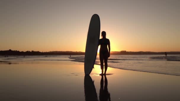 Щаслива молода жінка, що стоїть на піщаному пляжі, тримає дошку — стокове відео