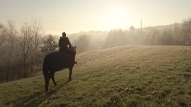Kadın sisli bir kış sabahı çayırda kahverengi bir ata biniyor..