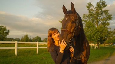 Gülümseyen genç bir kadın atını okşarken kameraya bakıyor..