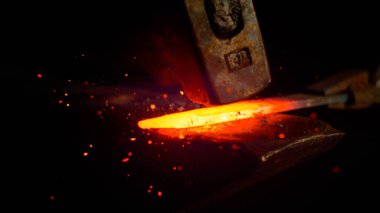 MACRO: Red hot piece of metal is held by tongs and struck by a big hammer.