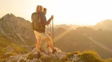 LENS FLARE: Unrecognizable cheerful hikers observe the golden sunset above Alps.