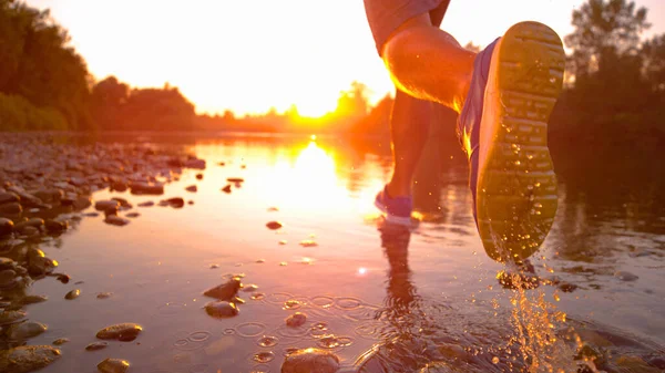 LENS FLARE, ZAMKNIJ nierozpoznany sportowiec jogging w płytkiej rzece o zachodzie słońca — Zdjęcie stockowe