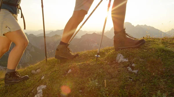 LENS FLARE: coppia turistica che indossa scarponi da trekking cammina su una collina erbosa nelle Alpi. — Foto Stock