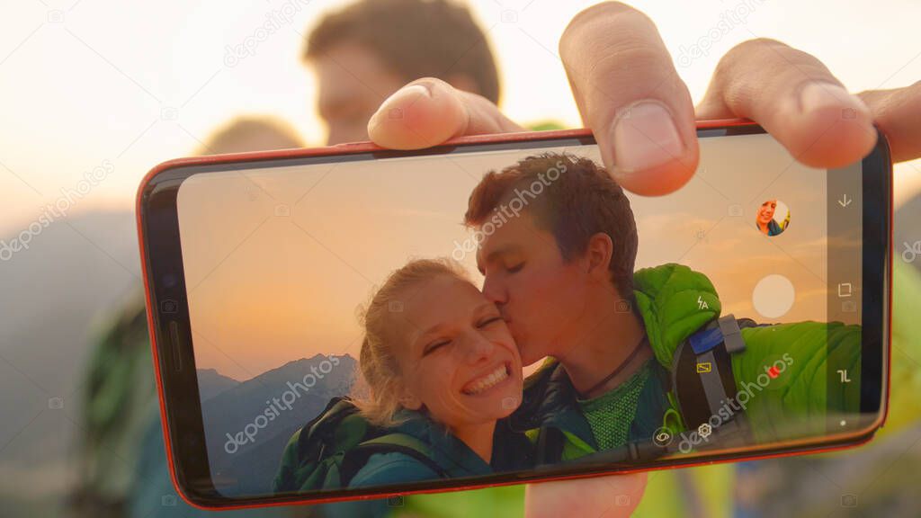 MACRO: Cheerful man takes a selfie of him kissing girlfriend on cheek at sunset.