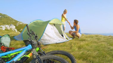 Smiling Caucasian woman hands a pin to her boyfriend setting up a pop up tent.