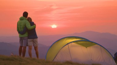 Unrecognizable man and woman stand embraced and watch the sunset in the Alps.