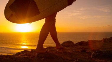 LOW ANGLE: Young woman walking and carrying surfboard along the rocky shore.