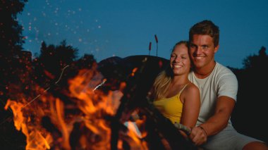 Kız arkadaşıyla kucaklaşan neşeli adam kamp ateşini bir sopayla dürtüyor..