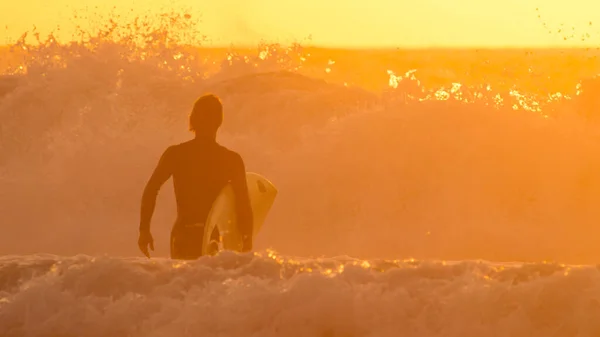 SILHOUETTE: Nierozpoznany surfer będzie surfować w oszałamiający złoty poranek. — Zdjęcie stockowe