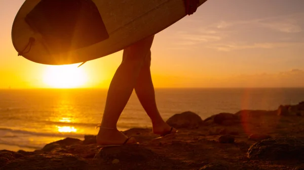 LOW ANGLE: Ung kvinna går och bär surfbräda längs den klippiga stranden. — Stockfoto