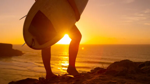 LENS FLARE: Girl holding a surfboard and observing the sunset from rocky hill. — ストック写真