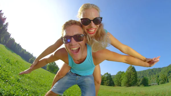 SUN FLARE: Homem animado dando a sua namorada um passeio de piggyback em um dia ensolarado. — Fotografia de Stock